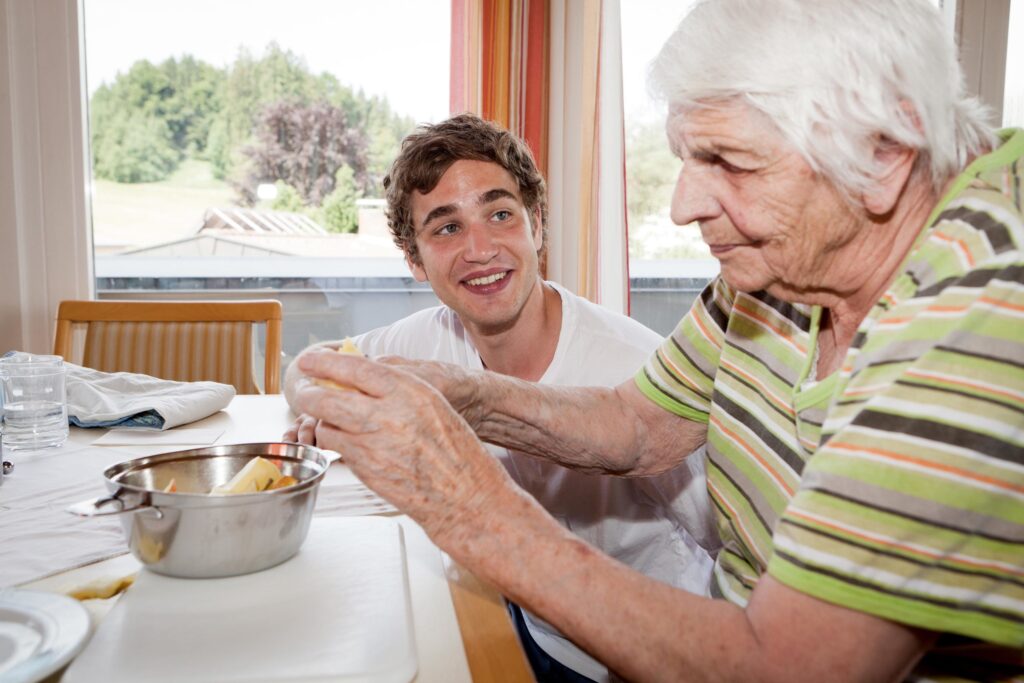 Pfleger kniet neben alter Frau, die am Tisch etwas backt